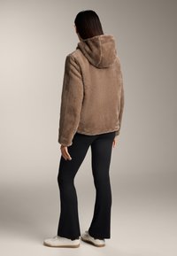 Brun, fluffig huva-jacka med avslappnad passform och långa ärmar, i kombination med svarta utsvängda byxor och vita sneakers. Slät bakgrund.