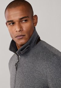 Chaqueta de mezcla de lana gris con cuello alto, cierre frontal con cremallera y botones a presión. Tejido texturizado con detalles en cuero marrón en el cuello.