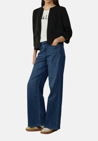 Zwarte cropped blazer, witte graphic tee en wijdvallende blauwe spijkerbroek. Beige en bruine sneakers met zijstrepen. Gladde texturen overal.
