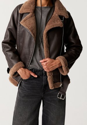 Personne portant une veste en cuir marron foncé avec une doublure en shearling beige, chemise grise et jean noir, tenant la veste partiellement ouverte.