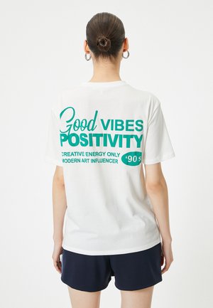 Hvid bomulds T-shirt med stor teal tekst på ryggen: "Good Vibes" og "Positivity" med yderligere sætninger i mindre skrift. Korte ærmer.