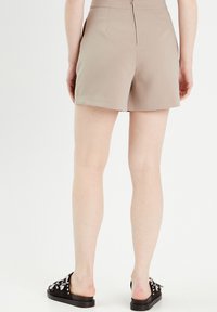 Pantaloni corti beige a vita alta con una texture liscia e una zip sul retro. Indossati con sandali neri slip-on con elementi metallici decorativi.