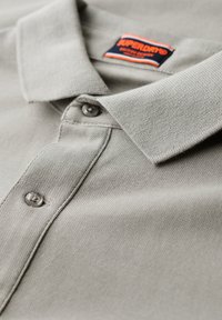 Lichtgrijze polo met een textuurstof, een kraag, twee knopen en een contrasterend merklabel dat in de kraag is gestikt.