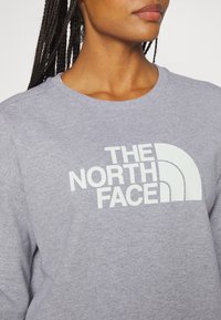 Sweat-shirt gris à col rond, avec un grand logo blanc "THE NORTH FACE" et des accents de design incurvés sur la poitrine.
