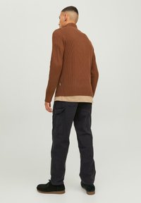 Suéter marrón de canalé con cuello alto, superpuesto a una camisa beige, combinado con pantalones cargo negros y botas de ante oscuras hasta el tobillo.