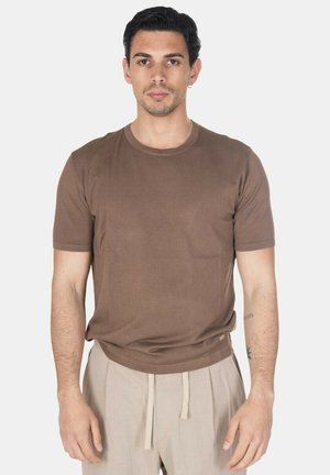 Uomo con capelli corti e scuri e orecchino, indossa una camicia marrone a maniche corte e pantaloni beige con coulisse, in piedi davanti a uno sfondo semplice.