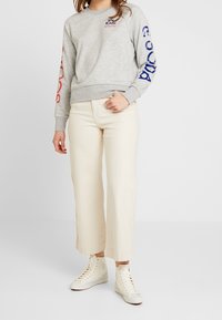 Sudadera gris con estampados azules y rojos en las mangas, combinada con pantalones anchos color crema y zapatillas blancas de caña alta. Estilo casual y cómodo.