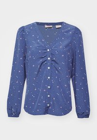 Blouse bleue à manches longues avec boutons blancs, détail froncé à l'avant et petit motif floral rose et blanc.