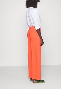 Pantaloni arancioni a gamba larga con vita cinturata e tasche posteriori, abbinati a una camicia bianca di lino a maniche corte e sandali marroni.