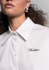 Weißes Hemd mit Kragen und Knopfschluss, das auf der linken Seite den schwarzen gestickten Text "Wrstbhvr" aufweist. Glattes Material und strukturiertes Design.