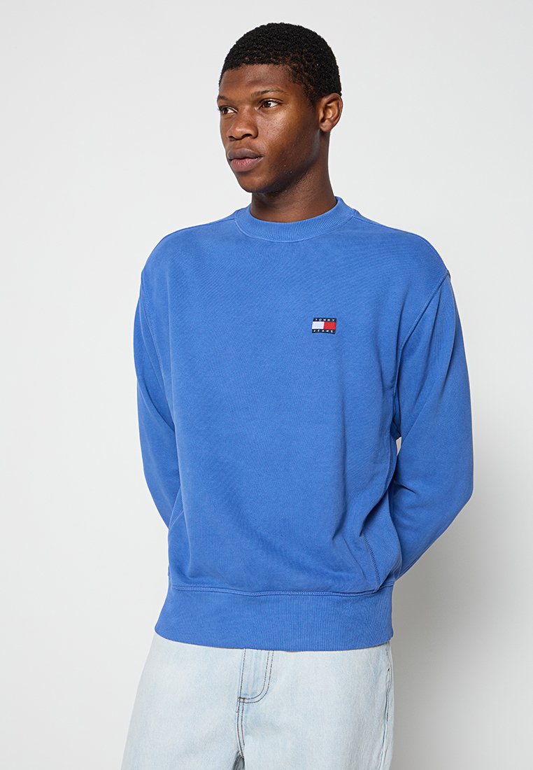 Niebieski bawełniany sweter z okrągłym dekoltem, ściągaczami przy mankietach i u dołu, z małą naszywką logo w czerwonym, białym i niebieskim kolorze na klatce piersiowej.
