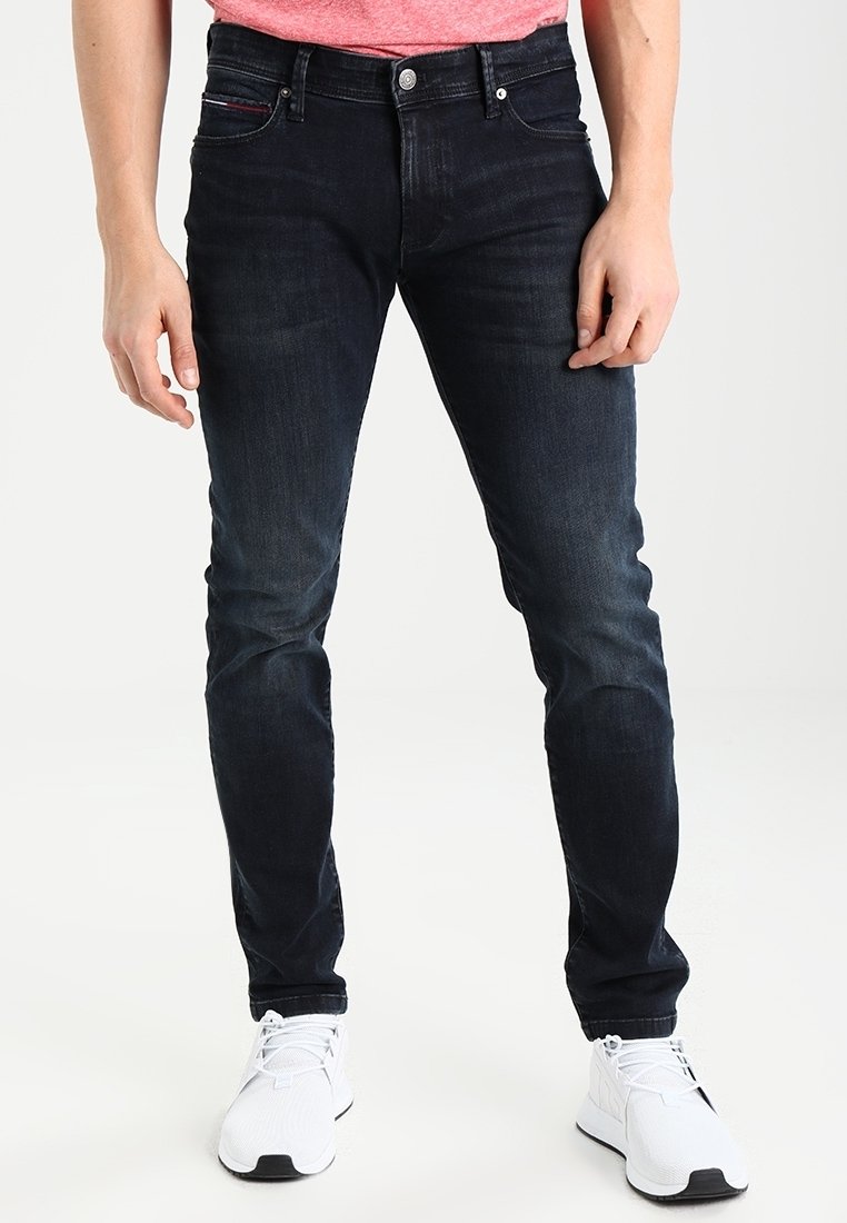 Homme portant un jean skinny foncé, des baskets blanches et une chemise rouge, debout devant un fond clair uni.