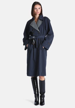 Femme portant un trench-coat bleu marine ceinturé avec de gros boutons et des bottes noires texturées montant jusqu'aux genoux, debout les mains dans les poches.