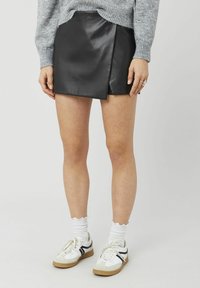 Mini-jupe en cuir noir avec un design portefeuille, associée à des chaussettes blanches à volants et des baskets beige et blanc. Le pull possède une texture tricotée ajustée.