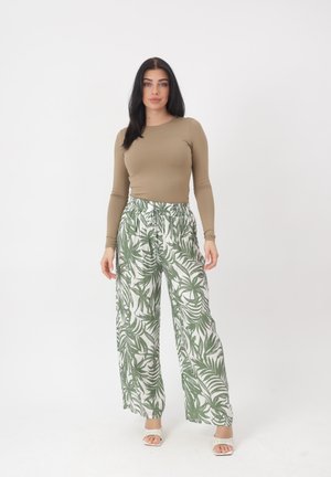 Frau mit langen dunklen Haaren, die ein beigefarbenes Langarmshirt, weiße Hosen mit weitem Bein und grünem Blattmuster sowie weiße Sandalen trägt und steht.