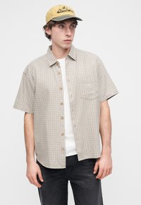 Πουκάμισο με κοντά μανίκια, μπεζ καρό, με ξύλινα κουμπιά και μία τσέπη στο στήθος, συνδυασμένο με κίτρινο καπέλο που φέρει μαύρο κεντημένο λογότυπο.