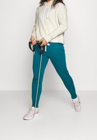 Felpa beige chiaro con tasca frontale e cordini bianchi; pantaloni jogger teal con tasche laterali; tiene delle bande di resistenza; indossa scarpe da ginnastica rosa chiaro.
