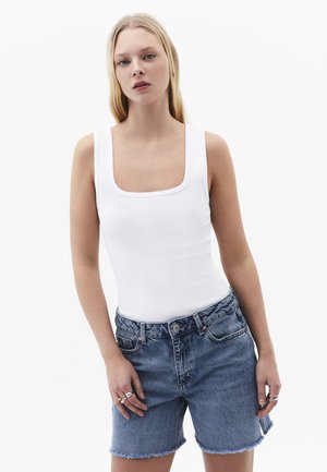 Femme portant un débardeur blanc côtelé sans manches et un short en jean bleu taille haute avec des bords effilochés, debout devant un fond blanc uni.