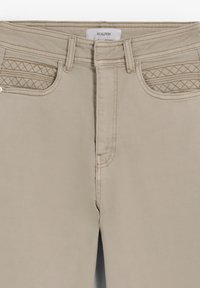 Pantalones beige con detalles cosidos en los bolsillos, cierre de botones y bragueta con cremallera. Fabricados con un tejido suave y texturizado. Etiqueta de la marca visible.