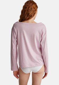 Persoon met lang, rood haar draagt een losse, lange mouwen roze top en witte ondergoed, met de rug naar de camera gekeerd tegen een eenvoudige witte achtergrond.