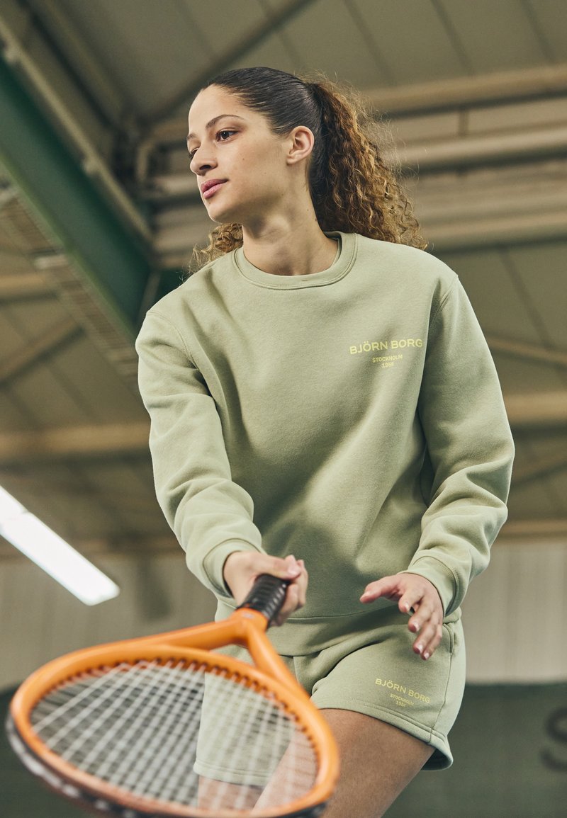 Felpa leggera verde chiaro e pantaloni corti con vestibilità ampia. Materiale in cotone, logo ricamato in giallo. Tiene in mano una racchetta da tennis arancione.