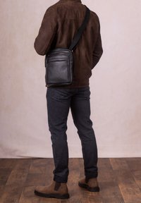 Black leather crossbody bag with a textured finish, featuring a front zipper, worn over a brown suede jacket with a black strap.