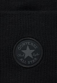 Material textil tricotat negru cu o etichetă circulară neagră care prezintă o stea și inscripția "Converse Chuck Taylor All Star" brodată cu fir negru.