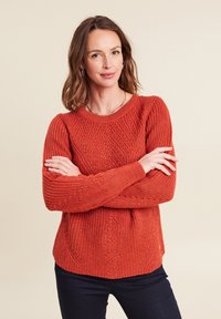 Maglione a maglia in arancione bruciato, con motivi testurizzati, colletto rotondo e maniche lunghe. Abbinato a pantaloni scuri.