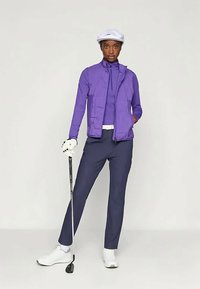 Γυναίκα που στέκεται με ένα μπαστούνι του γκολφ, φορώντας ένα λευκό καπέλο, μωβ σακάκι και μπλούζα, ναυτικές παντελόνες, λευκή ζώνη, λευκό γάντι του γκολφ και λευκά παπούτσια.