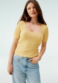Gul ribbad kortärmad topp med fyrkantig halsringning, i kombination med ljusblå högmidjade jeans. Slät textur och åtsittande design.
