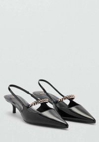 Svarta lackläders slingback-klackar med spetsiga tår, smala remmar och en guldig kedjeaccent över fotvalvet. Lågt, staplat klack.