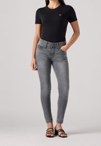 Svart bomulls-T-shirt med en liten röd logotyp, kombinerad med grå tajta jeans med en klassisk femfickdesign och en lätt blekning. Svarta sandaler.