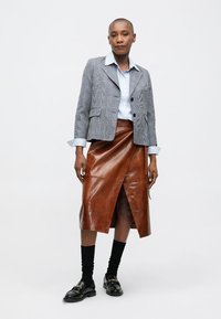 Γυναίκα, που στέκεται, φορώντας ένα γκρι καρό μπλέιζερ, ανοιχτό μπλε πουκάμισο, καφέ δερμάτινη μίντι φούστα με πλαϊνό σκίσιμο, μαύρες κάλτσες και μαύρες loafers με χρυσές λεπτομέρειες.