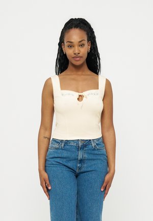 Femme aux cheveux longs tressés portant un haut sans manches crème avec une petite broderie florale bleue et un jean bleu taille haute, de face.