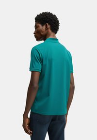 Polo shirt turchese con colletto e maniche corte, caratterizzato da una texture liscia. Il modello indossa jeans scuri, visto di spalle, rivolto a sinistra.