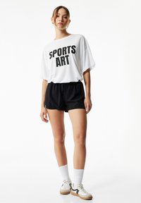 Biała, oversize'owa koszulka z napisem "SPORTS ART" w pogrubionym czarnym tekście, połączona z czarnymi spodniami sportowymi i białymi skarpetkami do kolan.