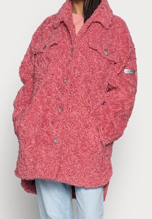 Veste surdimensionnée rose texturée avec boutons et poches poitrine, portée par-dessus un haut rose clair et un jean bleu clair.