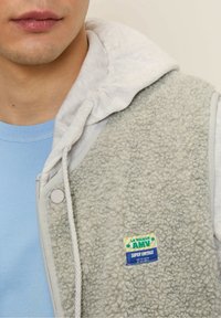Gray fleece vest with a white hoodie, featuring a blue label that reads "AMY SUPER VINTAGE," worn over a light blue shirt.