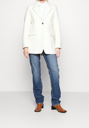 Personne portant un blazer blanc texturé, une chemise blanche à col, un jean bleu et des bottines marron à bout carré, debout devant un fond uni.