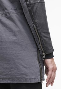 Lång grå kappa med en strukturerad, tygliknande kropp och kontrasterande läderärmar. Har sidofickor med dragkedja och ribbade muddar, vilket förbättrar rörligheten.