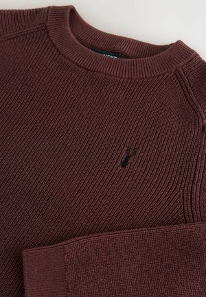 Burgundská pletená mikina s rebrovanou textúrou, okrúhlym výstrihom a malým čiernym logom na hrudi. Rukávy majú voľný strih.