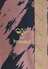Hummel sportkläder med en rosa bas och mönster av marinblå och svarta penseldrag, med en gyllene broderad logga och guldiga chevron-accenter.