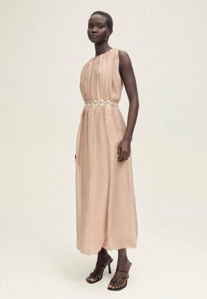 Robe longue sans manches de couleur rose pâle, fabriquée en tissu léger, avec des détails froncés et une ceinture circulaire métallique. Talons noirs à lanières.