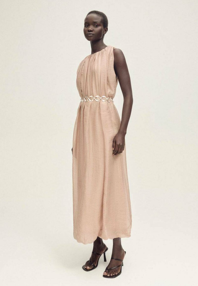 Robe longue sans manches de couleur rose pâle, fabriquée en tissu léger, avec des détails froncés et une ceinture circulaire métallique. Talons noirs à lanières.