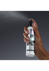 Bouteille pulvérisateur blanche tenue à la main, avec un bec pulvérisateur noir et un texte multicolore. Les étiquettes indiquent "Unis par un même but" et "25 avantages".