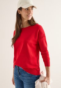 Rood wollen trui met lange mouwen en een ronde hals, gecombineerd met blauwe jeans. De textuur lijkt zacht; er wordt een lichtgekleurde pet gedragen.