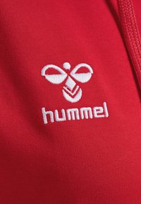 Roter Baumwollstoff mit einem weißen gestickten Logo, das stilisierte Formen zeigt, und dem Wort "hummel" darunter. Glatte Textur.