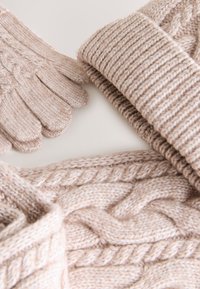 Light beige knit hat, gloves, and scarf. Features textured patterns and cables. Soft, thick material with ribbed and woven details.