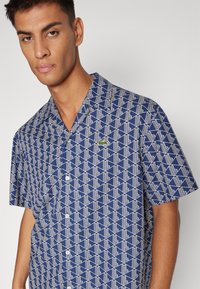 Camisa de botones de manga corta en color marino con un patrón geométrico blanco y un pequeño logo verde en el pecho. Tejido ligero, corte holgado.