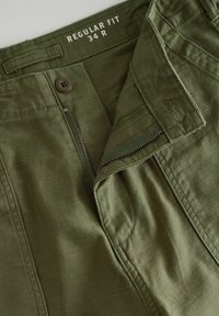 Pantalons vert olive à coupe régulière avec fermeture à bouton, braguette zippée et poches cousues. Le tissu semble lisse avec un léger éclat.
